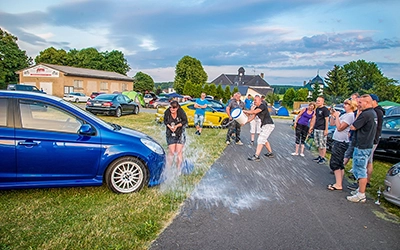 Rennstrecke Schleizer Dreicke Nach einer Siegerehrung bei einem Motorsport-Treffen wird eine Person humorvoll mit einem Eimer Wasser bespritzt, während andere Zuschauer lachen und Autos im Hintergrund geparkt sind. Aufgenommen von Fotograf Michael Schalansky.
